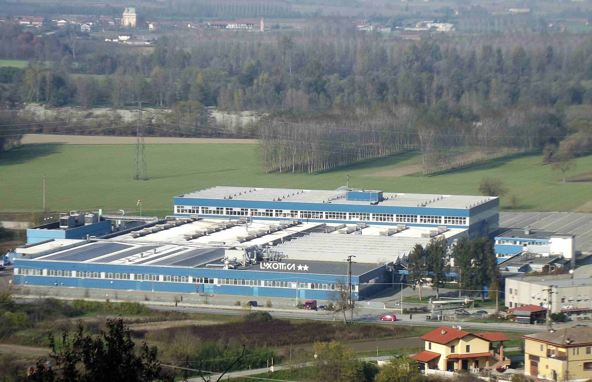 Veduta dall'alto dello stabilimento Luxottica di Lauriano, in provincia di Torino: capannoni azzurri e bianchi con l'insegna Luxottica sul tetto, circondati da campi e boschi.