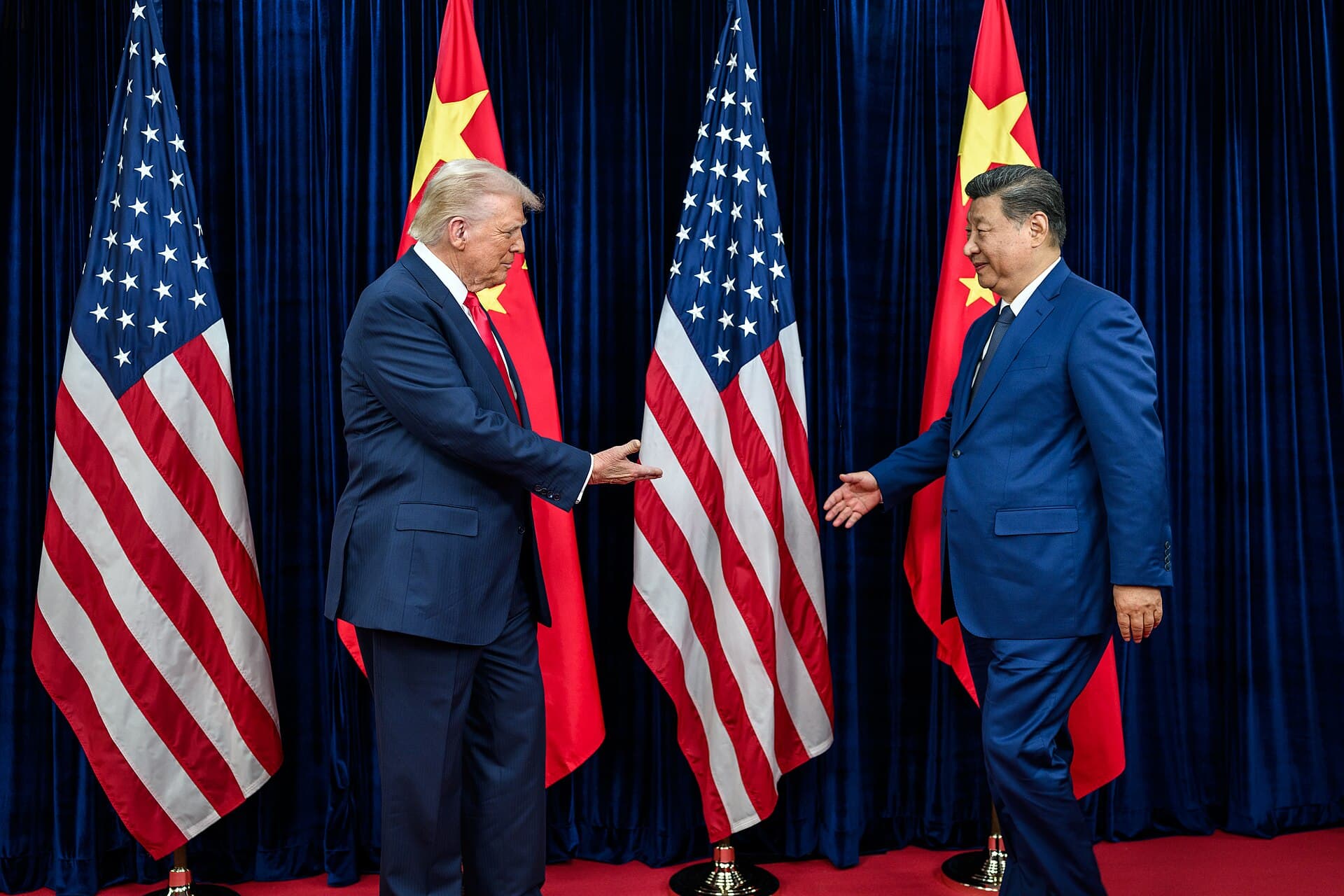 Donald Trump and Xi Jinping reach out to shake hands in front of the United States and China flags, against a backdrop of blue curtains, before their meeting in Busan on October 30, 2025