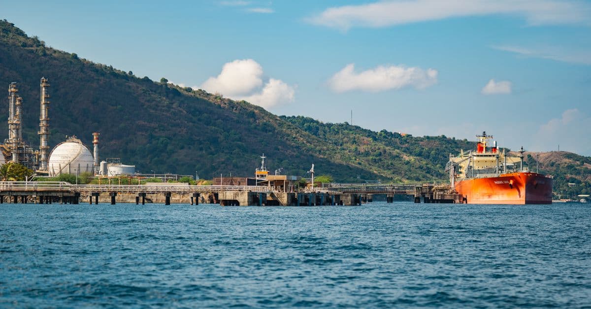 Una petroliera arancione ormeggiata al pontile di un terminal petrolifero costiero, con impianti di raffineria e colline verdi sullo sfondo
