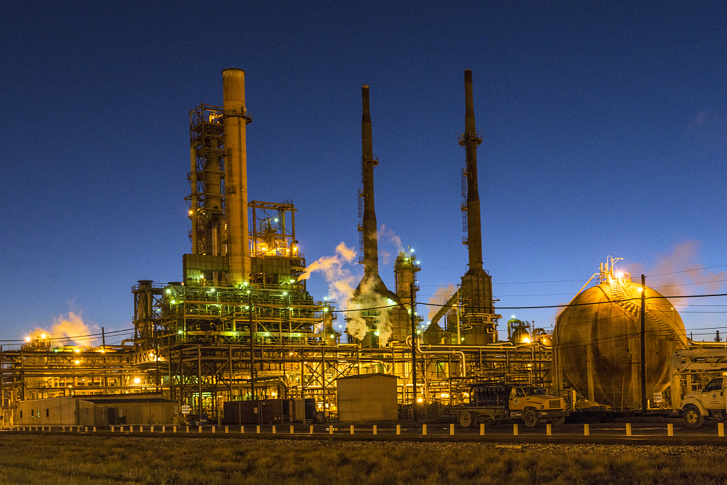 Una raffineria di petrolio illuminata al crepuscolo, con tre alte ciminiere, impianti accesi e un serbatoio sferico